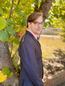 Derek stands in front of a tree wearing a dark grey suit, pink dress shirt, and fun black tie.  They're wearing their glasses and looking over the shoulder with their hair pulled over to the side.
