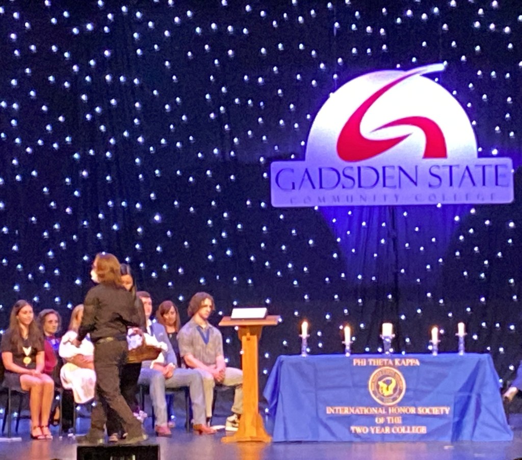 Derek walks with their back turned past a table with a purple Phi Theta Kappa tablecloth with lit candles and an outward facing podium. They have a white rose in their hand and are wearing all black.