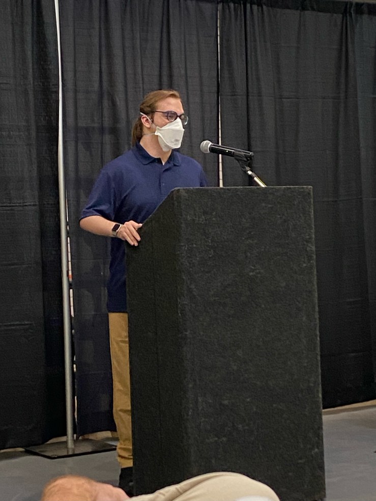 Derek stands at a podium delivering their speech. They are wearing blue glasses, a mask, a watch, a navy shirt, and khaki pants. Their hair is pulled back into a ponytail.