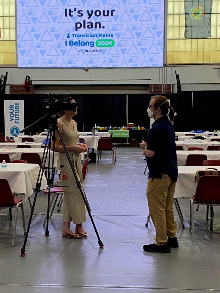 A photo of Derek talking to a reporter in the back of the auditorium. There is a camera positioned in front of them and they are looking at the reporter, Caroline, who is wearing a tan dress and holding papers. Derek is wearing blue glasses, a mask, a watch, a navy shirt, and khaki pants with their hair pulled into a ponytail.