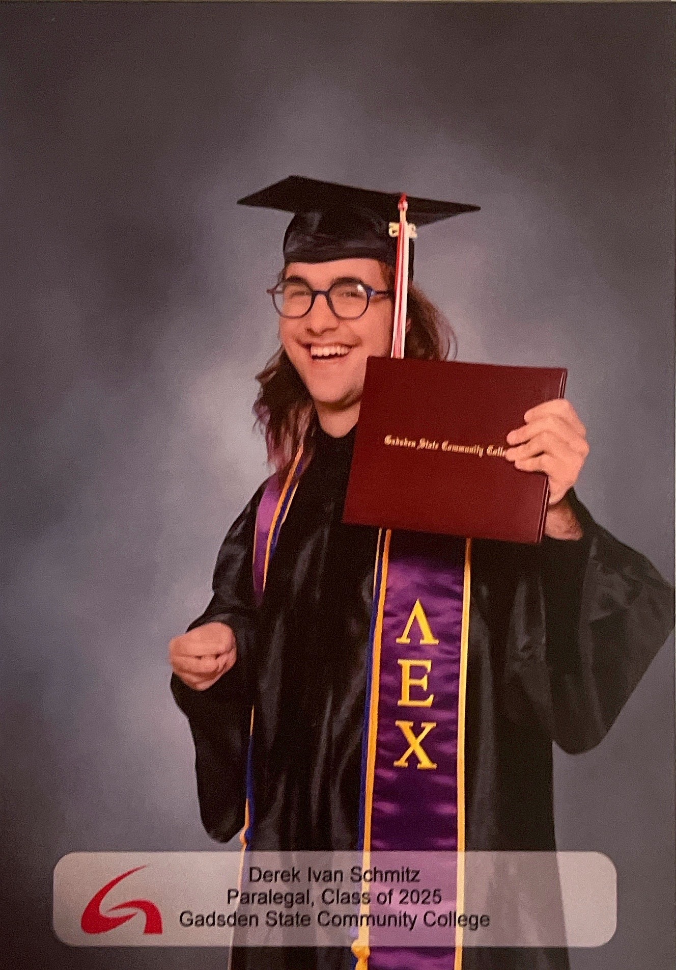 Derek, a white nonbinary person dressed in graduation regalia, including cap and gown, an honor chord, and Lambda Epsilon Chi honor society stole holds a diploma cover. 

A frosted textbox on the bottom of the image houses text that reads: 

Derek Ivan Schmitz
Paralegal, Class of 2025
Gadsden State Community College

as well as the Gadsden State initialmark - a stylized G.