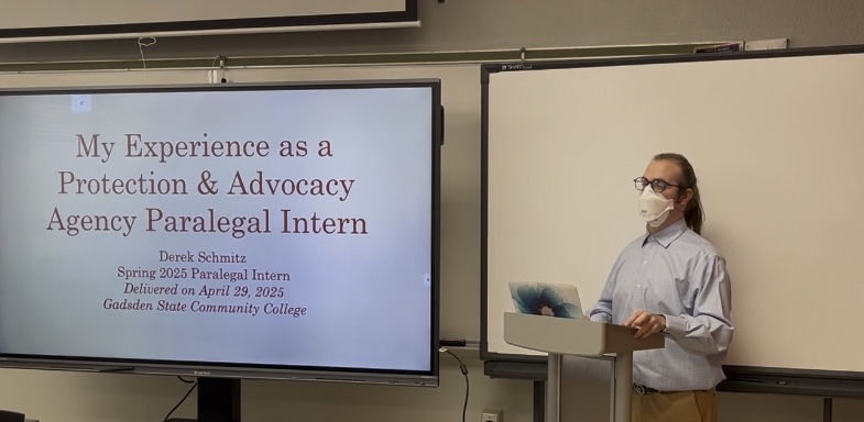 Derek, a white nonbinary person with shoulder-length brown hair in a ponytail, wearing a blue button-down shirt, khaki pants, and blue glasses stands behind a lectern. On a large screen to their left, a PowerPoint title slide bearing the words:

"My Experience as a Protection & Advocacy Agency Paralegal Intern

Derek Schmitz

Spring 2025 Paralegal Intern

Delivered on April 29, 2025

Gadsden State Community College"

displayed. Derek is turned to the audience, delivering the presentation.